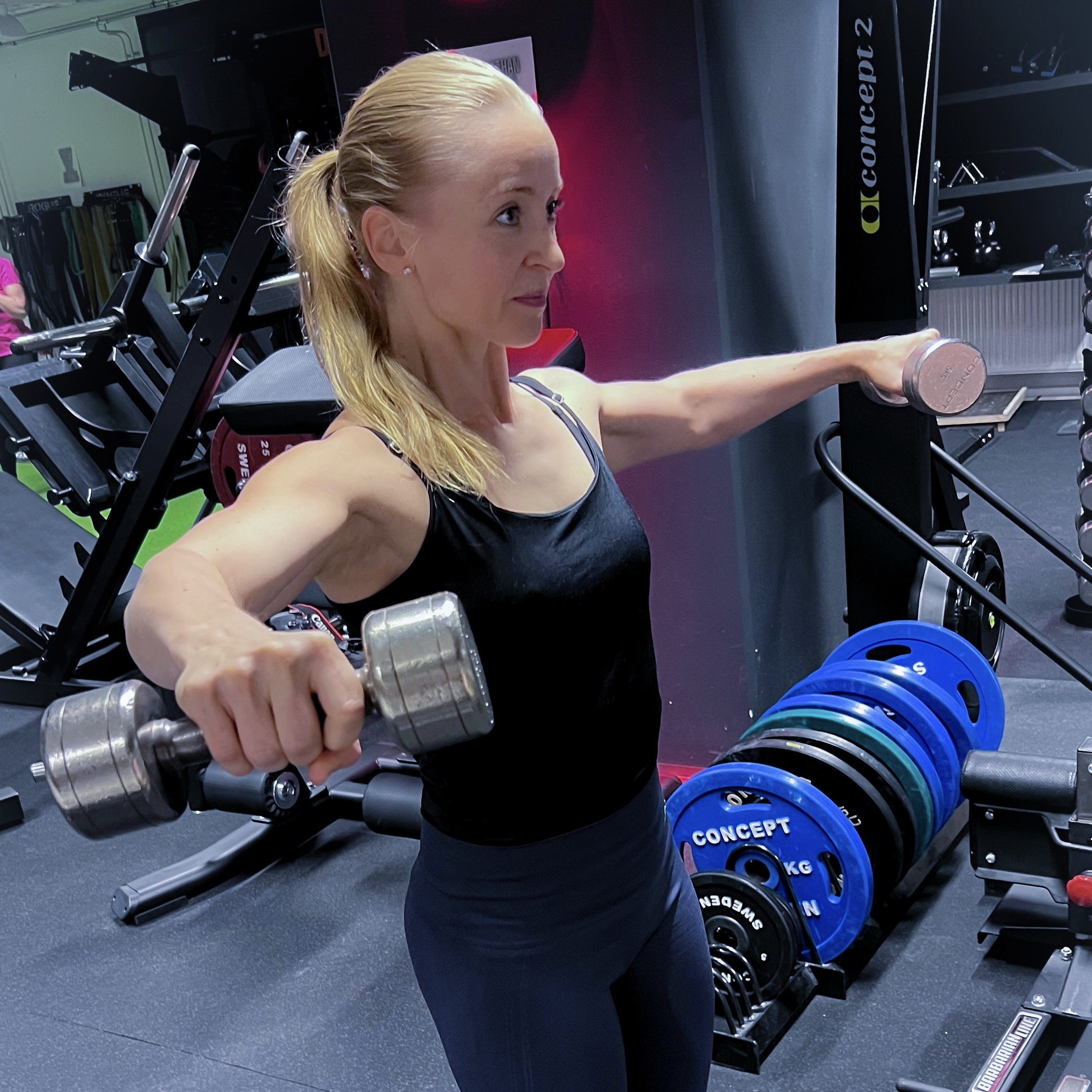 The trainer lifts two dumbbells in a front raise in a well-equipped gym