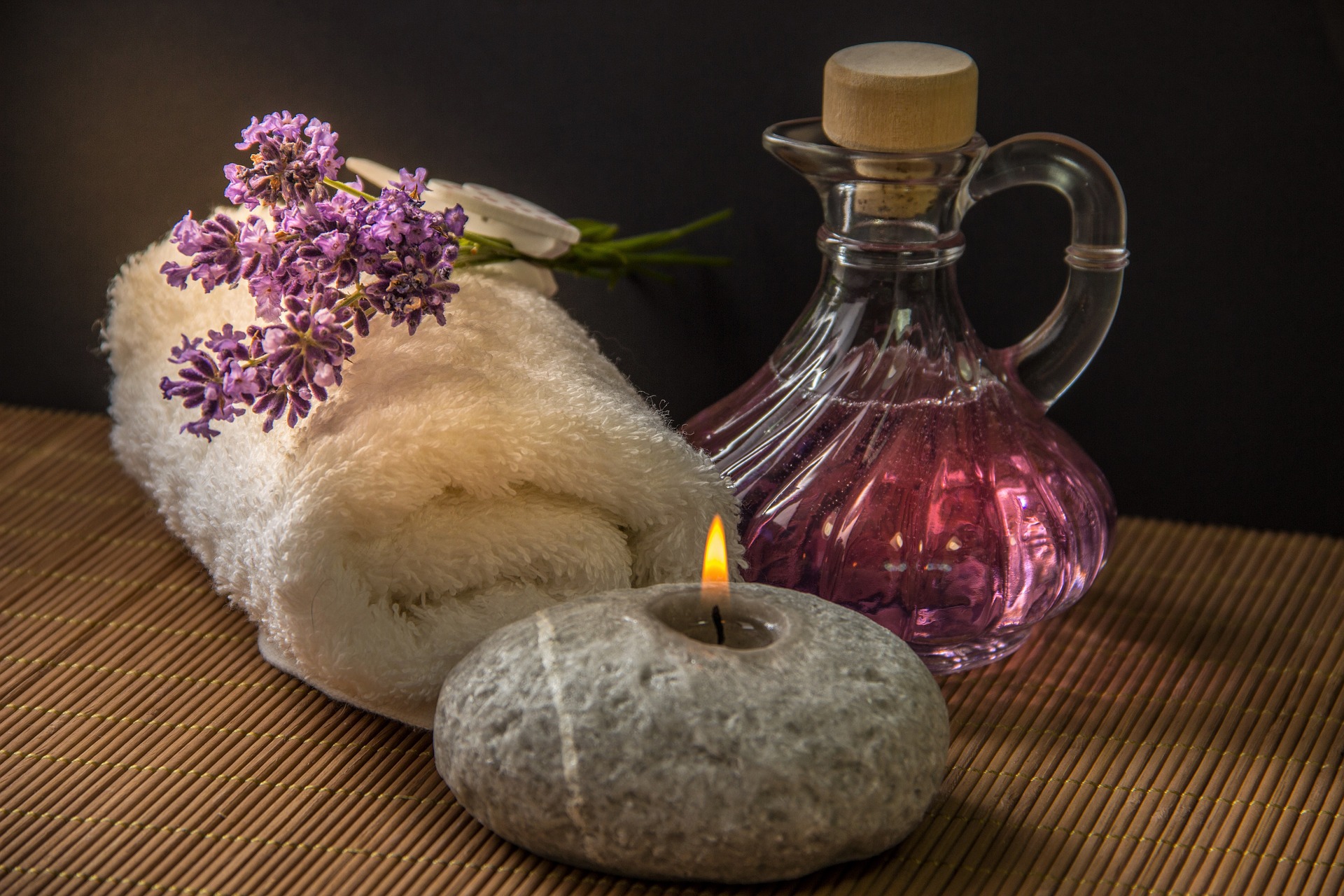 A rolled white towel with lavender, a jug of massage oil and a lit candle on a bamboo mat